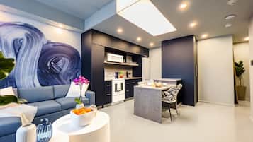 Una sala de estar moderna con un sofá, una mesa de centro y objetos decorativos. Se ve un área de cocina con una mesa de comedor y sillas al fondo.