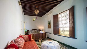 Quarto com chão xadrez, cama com roupa de cama estampada em vermelho e branco, um armário de madeira, uma pequena mesa redonda com toalha branca e uma janela com grades de metal.