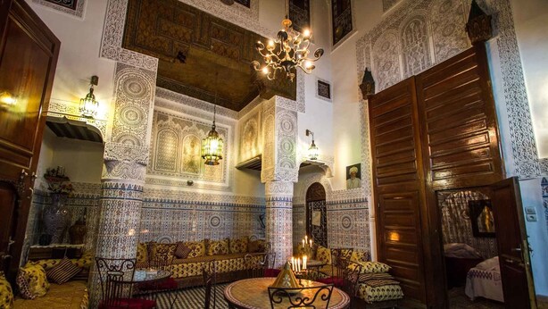 Um interior marroquino com trabalhos detalhados em azulejos, uma mesa redonda com cadeiras e uma grande porta de madeira.