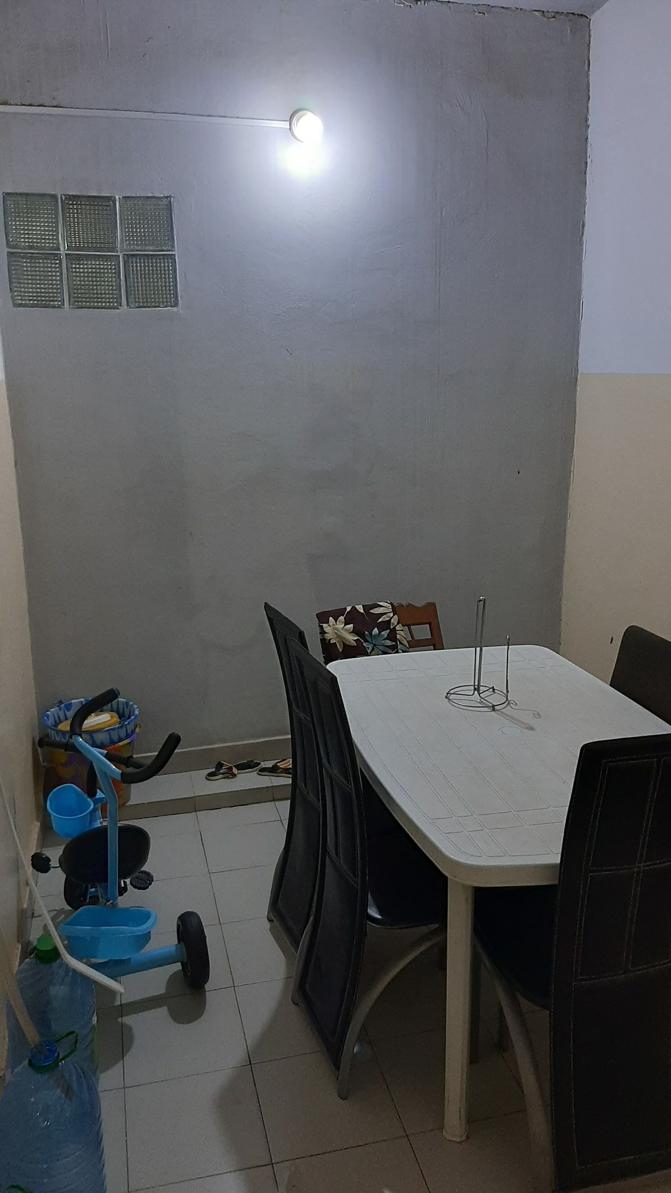 Une petite pièce peu éclairée, avec une table et des chaises blanches, un tricycle bleu et un seau bleu.