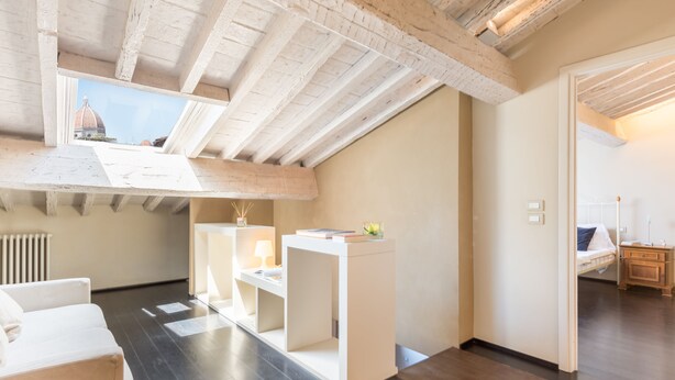 Een moderne loft slaapkamer met zichtbare houten balken, dakraam, een wit bed, bureau en uitzicht op een koepelgebouw.
