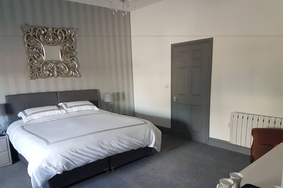 Quarto de hotel moderno com uma cama grande, um espelho decorativo, uma porta e uma pequena mesa com chávenas e um tabuleiro.