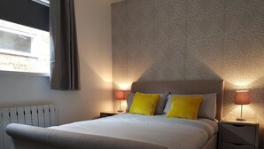A bedroom with a large bed, two bedside lamps, a radiator, and a window with curtains. - The Dalby Rooms (Margate)