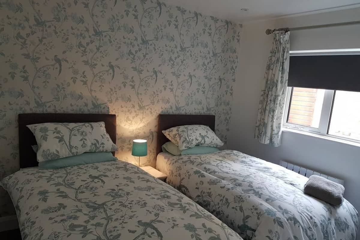 Quarto com duas camas, roupa de cama estampada com motivos florais, abajur e janela com cortinas.