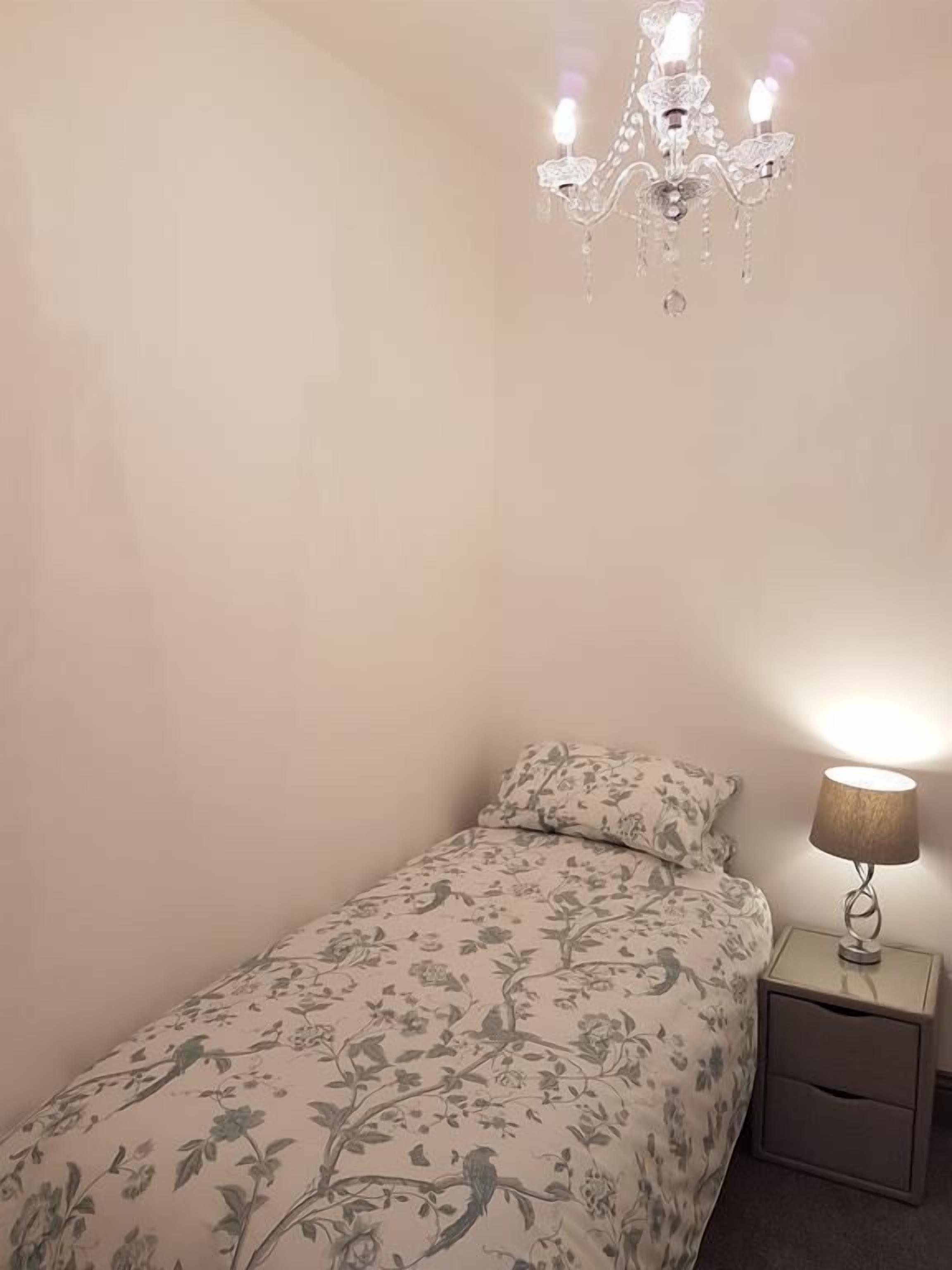 A small bedroom with a single bed, a nightstand, a lamp, and a chandelier.