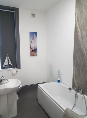 A bathroom with a white sink, a bathtub, and a window with a sailboat painting.