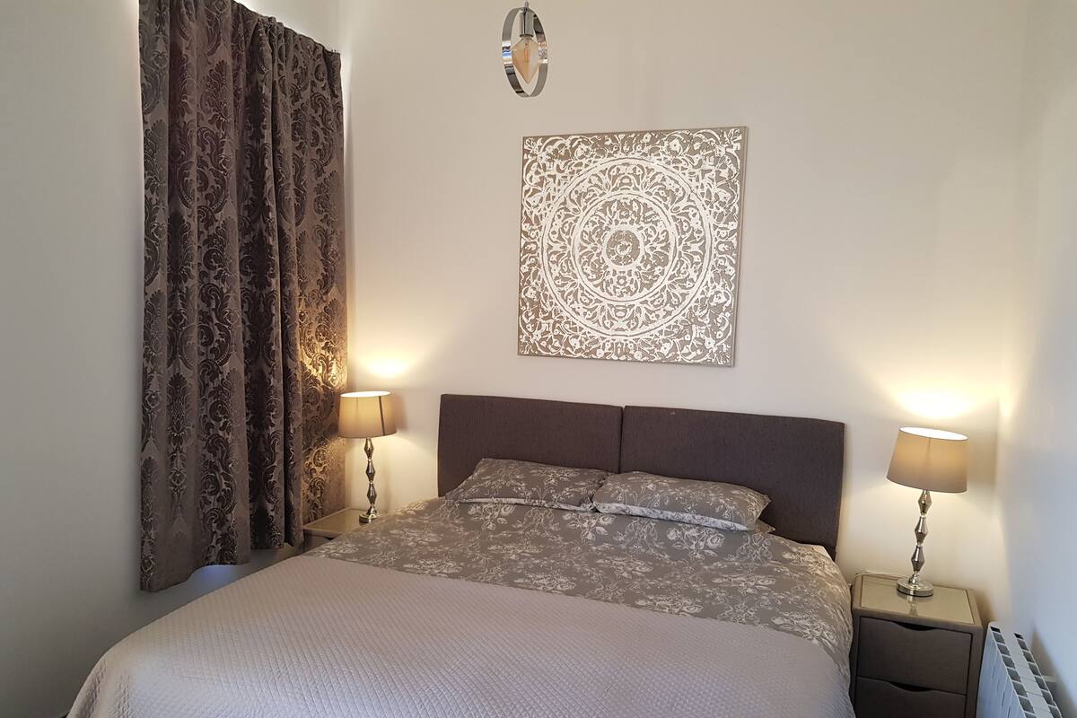 Quarto bem arrumado com uma cama grande, duas luminárias de cabeceira, um objeto decorativo na parede e um radiador.
