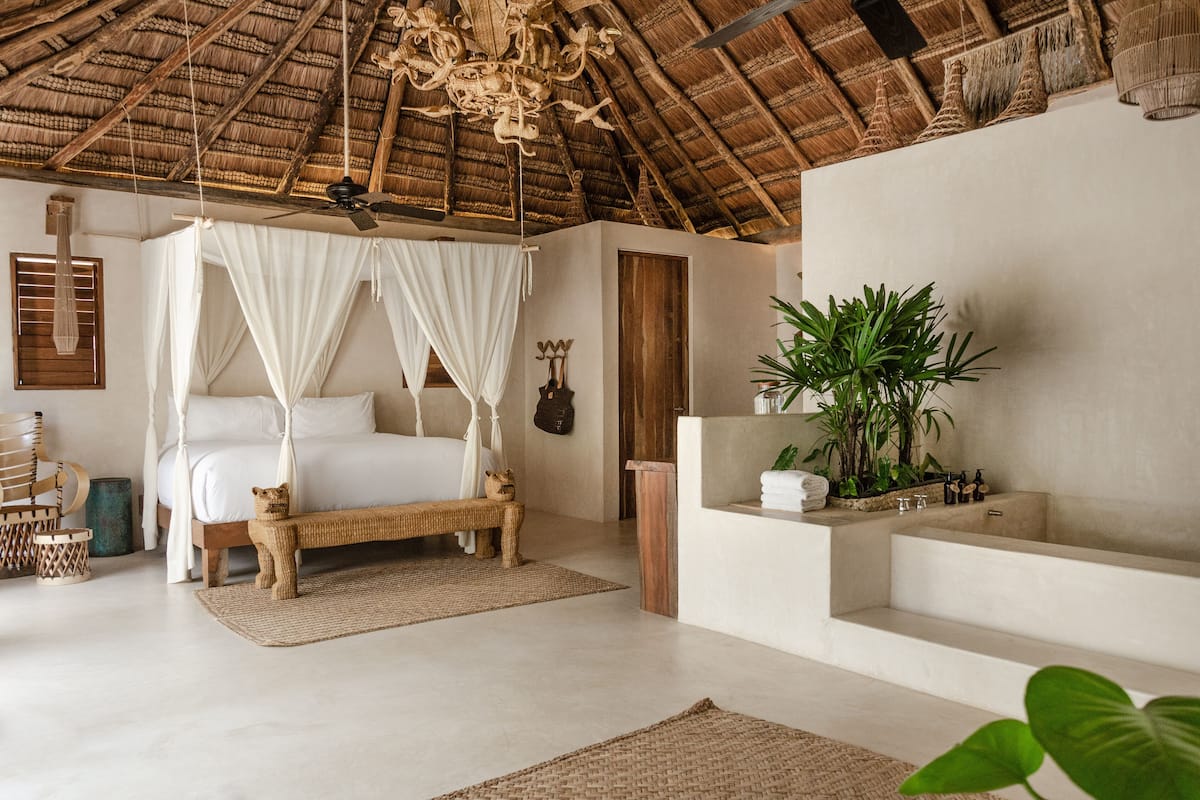 A bedroom with a four-poster bed, a wooden bench, a small cat statue, a plant, and a bathtub.