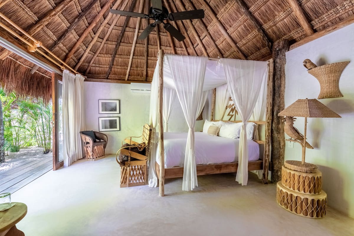 A room with a thatched roof, a bed with a canopy, a wooden chair, a woven table, and a wall-mounted lamp.