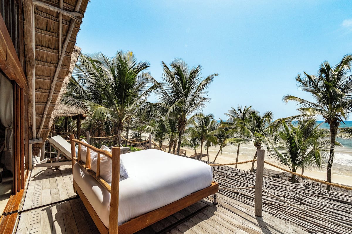 A wooden deck with a bed, palm trees, and a beach view.