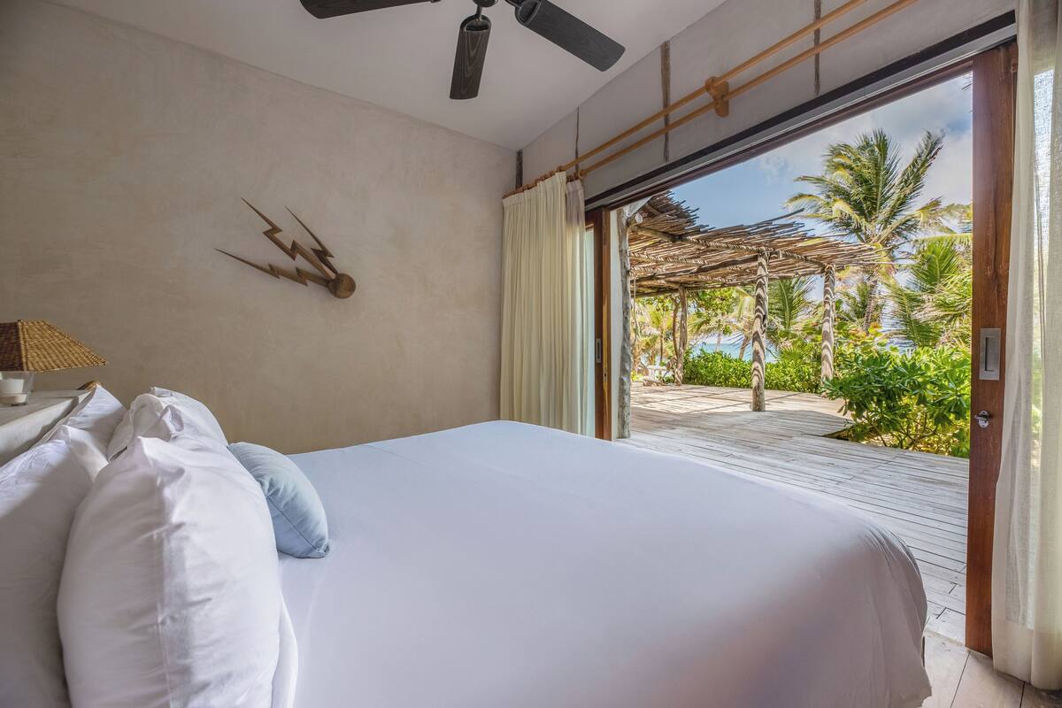A bedroom with a bed, a ceiling fan, a wall clock, and a view of a palm tree garden through a sliding door.