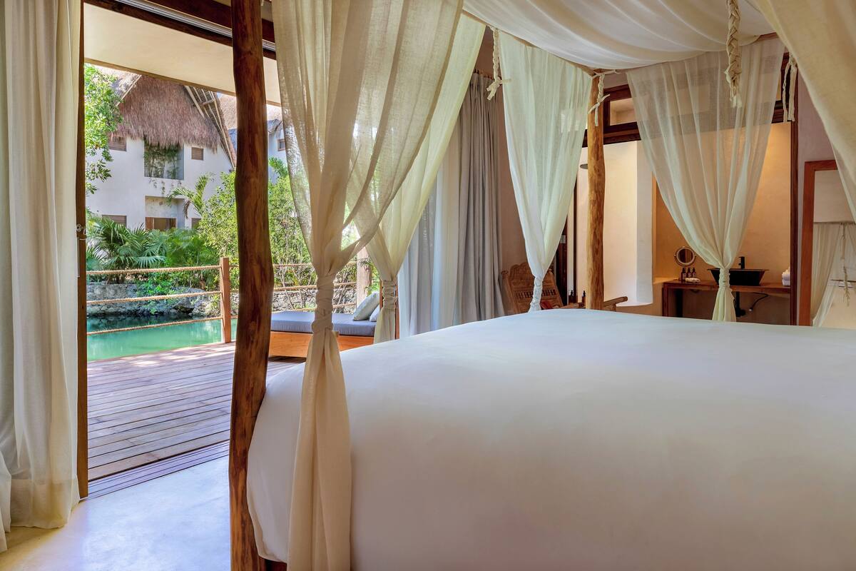 A bedroom with a large bed, white curtains, and a view of an outdoor pool and greenery.