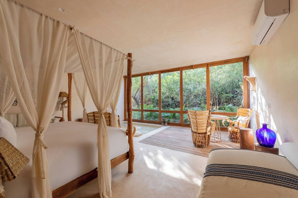 A bedroom with a four-poster bed, a wooden floor, and a view of the outdoors through large windows.