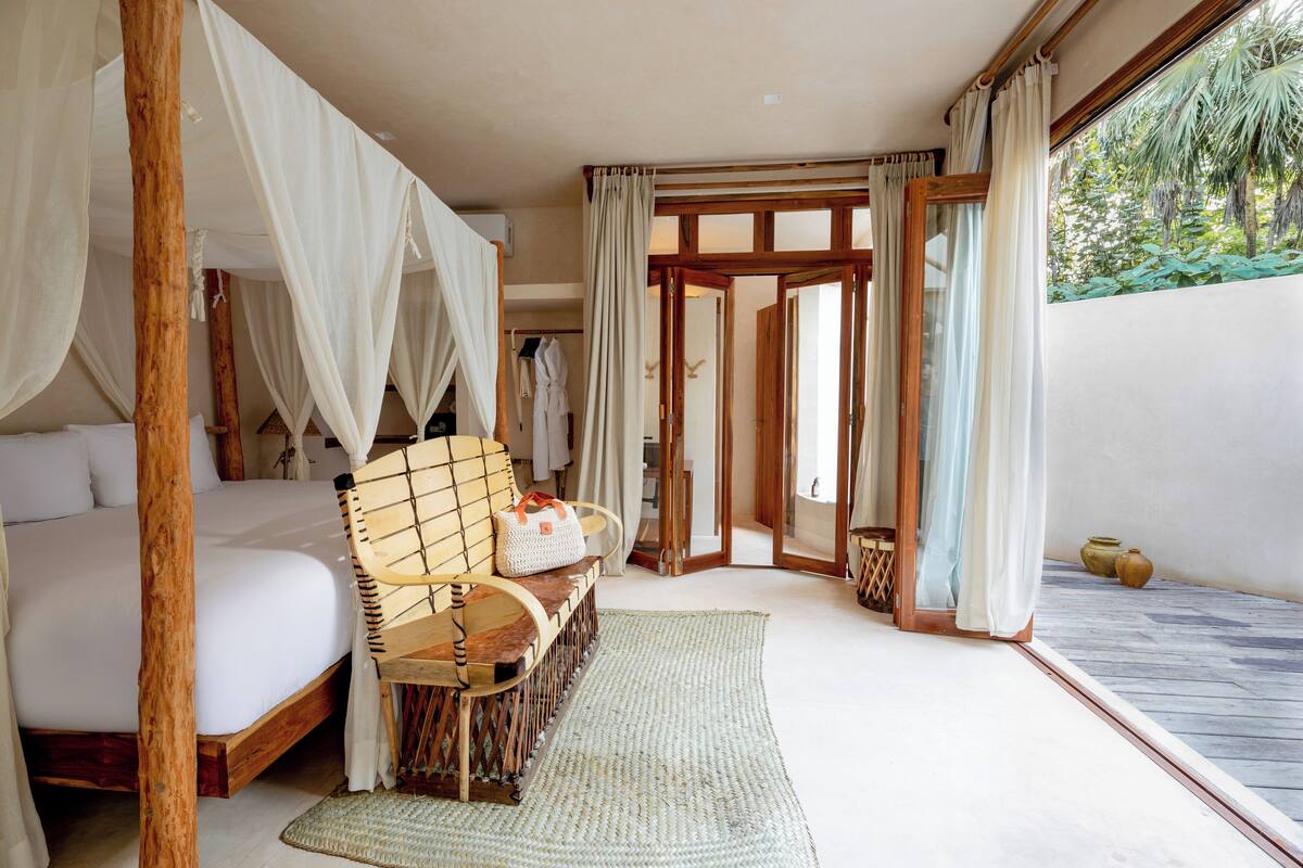 A bedroom with a four-poster bed, a wooden chair, a woven rug, and a view of the outdoors.
