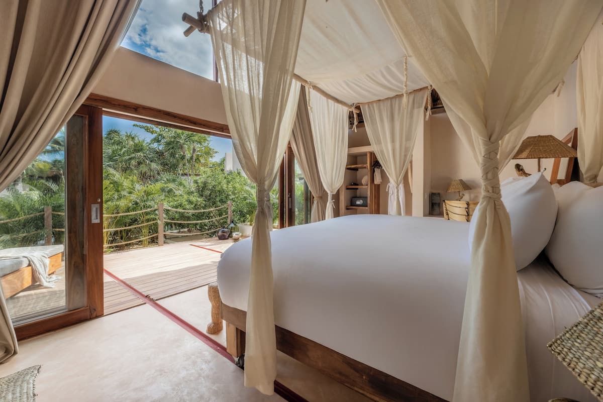 A bedroom with a large bed, a wooden floor, and a view of greenery outside.