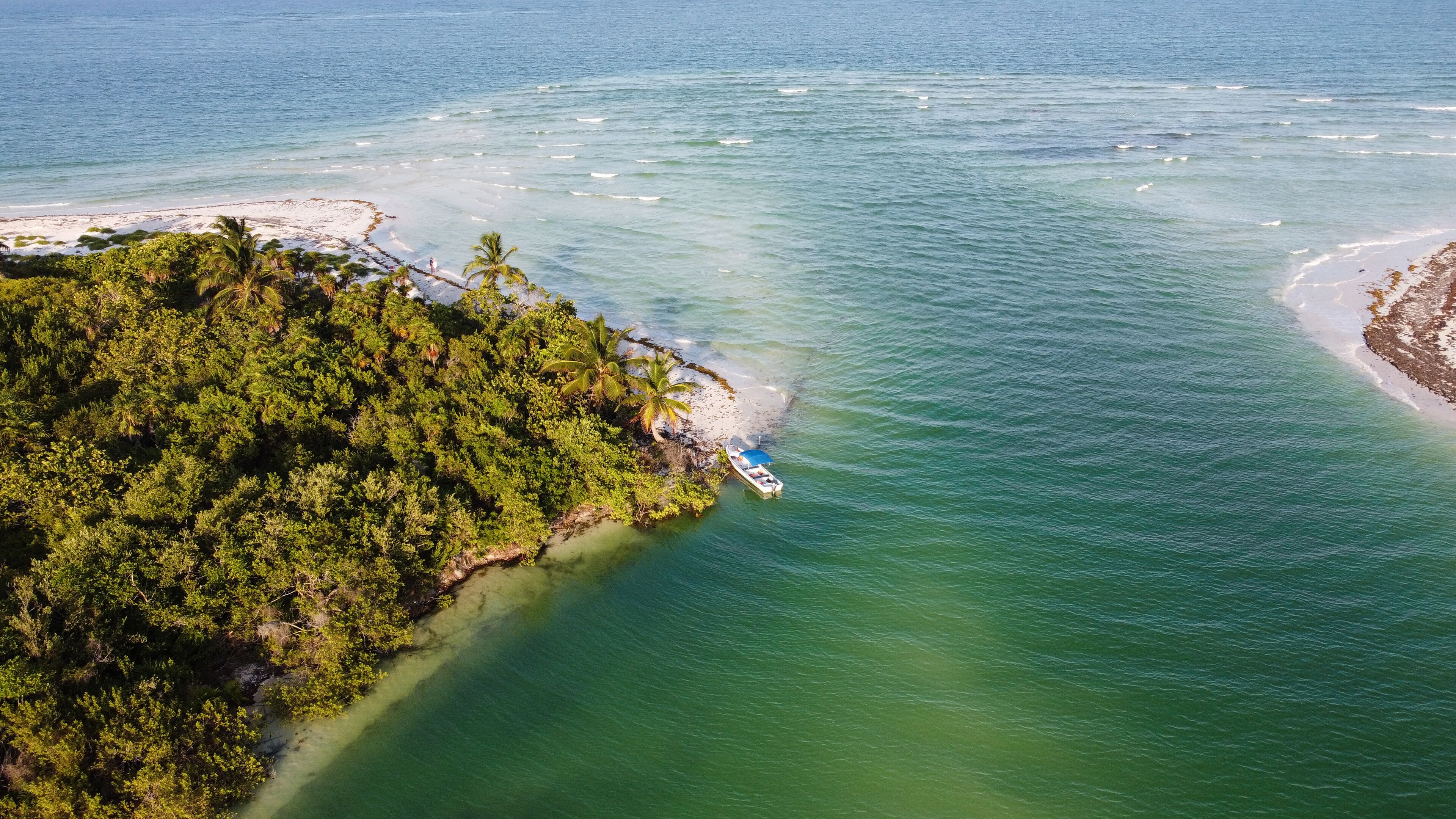 An aerial view of a tropical island with a boat docked near the shore, surrounded by clear waters and dense vegetation.