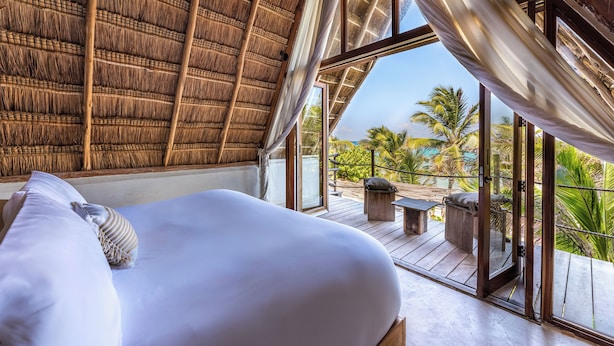 Una habitación con cama, ventanas amplias y vista a palmeras.