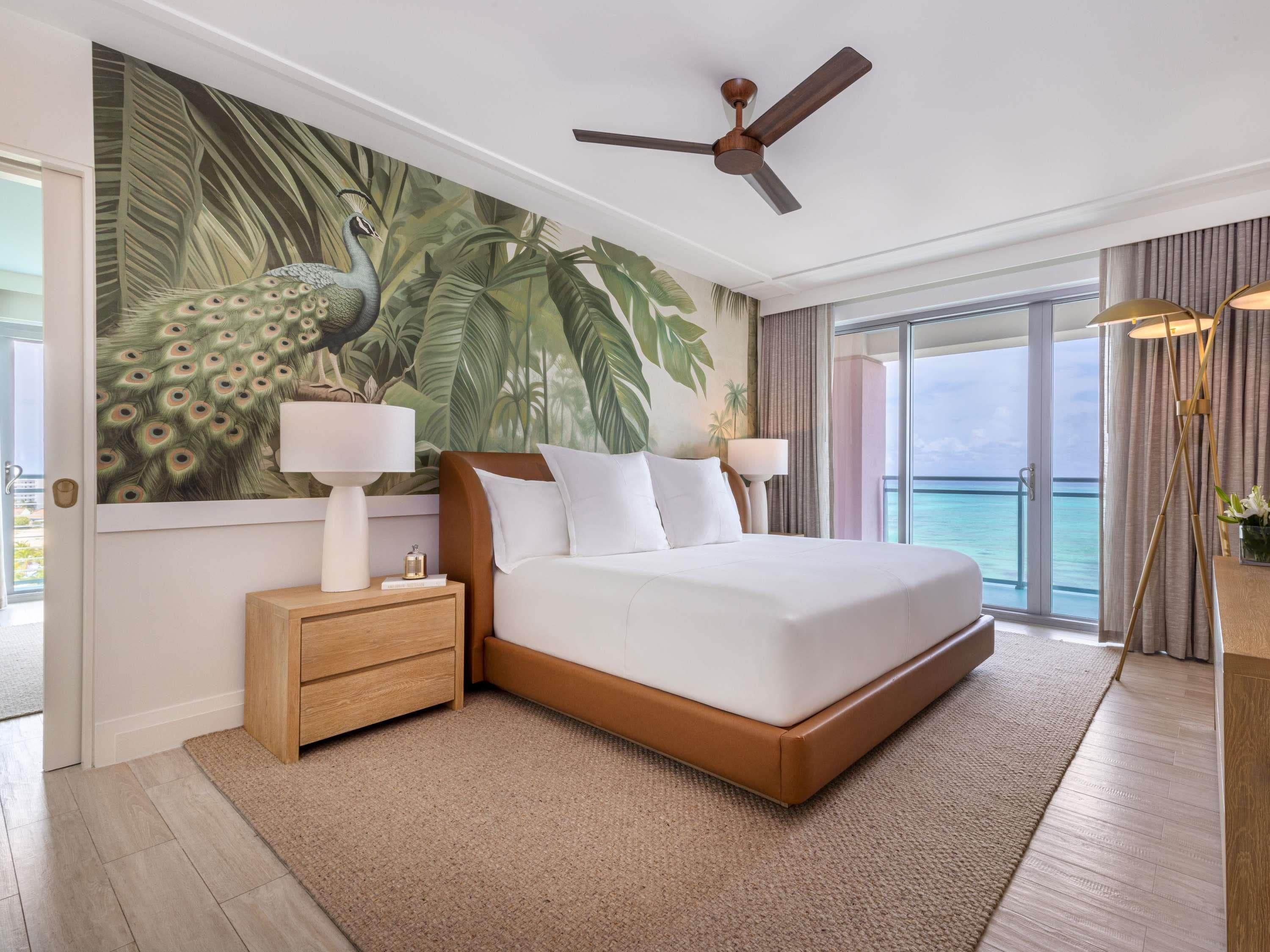A bedroom with a large peacock mural, a ceiling fan, a bed with white linens, a nightstand with a lamp, and a view of the ocean through large windows.