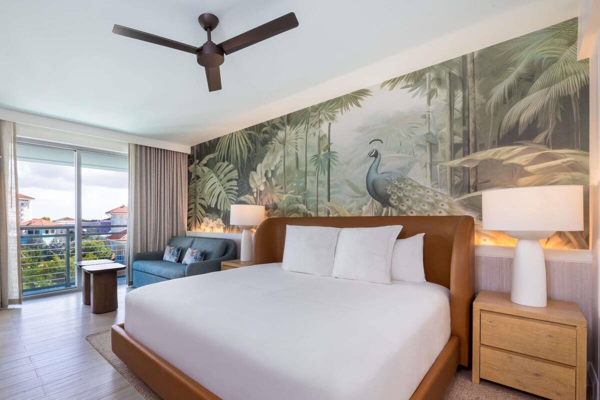 A hotel room with a large bed, two bedside tables, a ceiling fan, a view of the city, and a tropical mural.