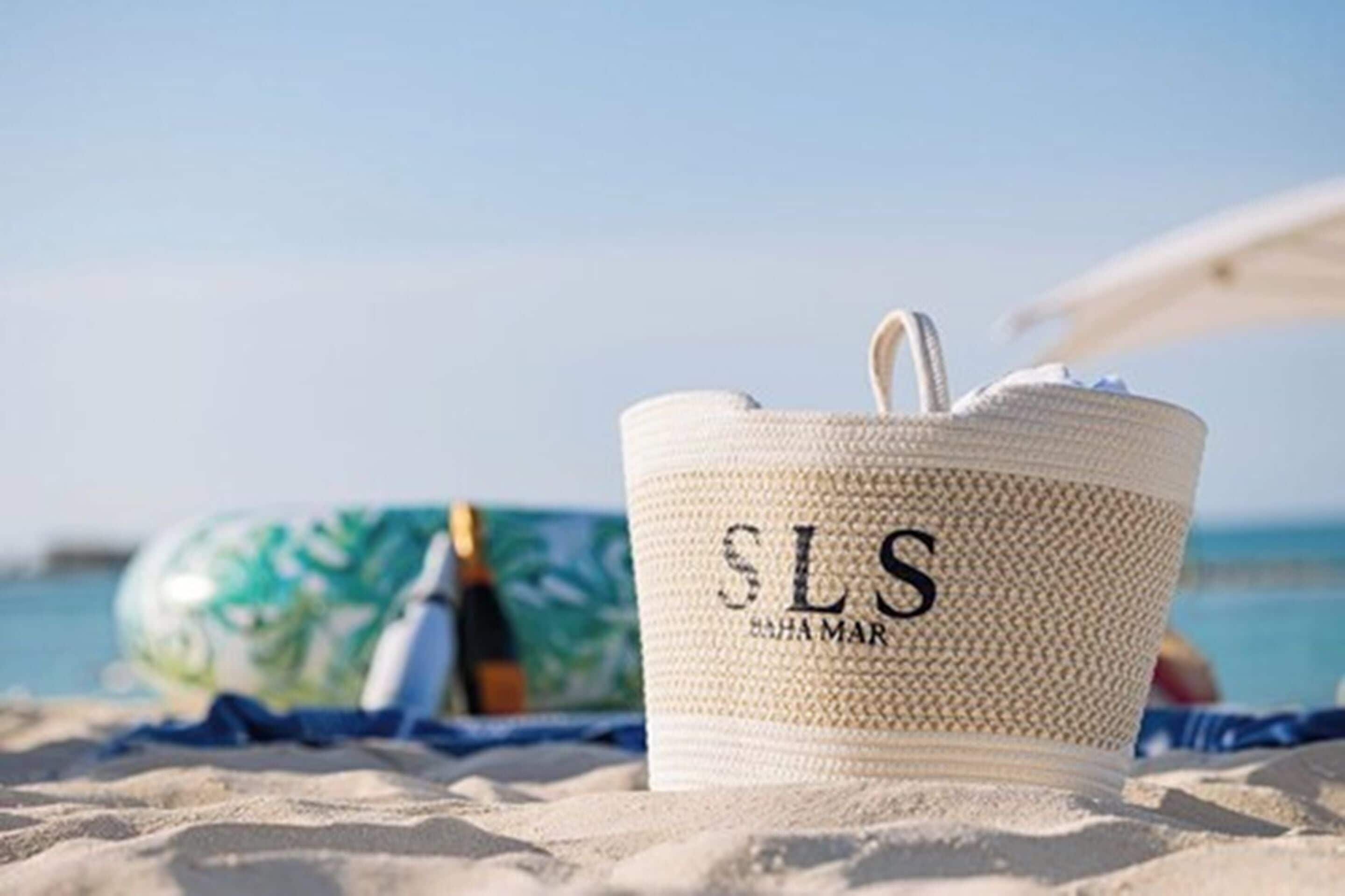 Una bolsa de playa blanca tejida con las iniciales 'SLS' y el texto 'SAHA MAR' sobre una playa arenosa, con un cielo azul despejado y el océano al fondo.