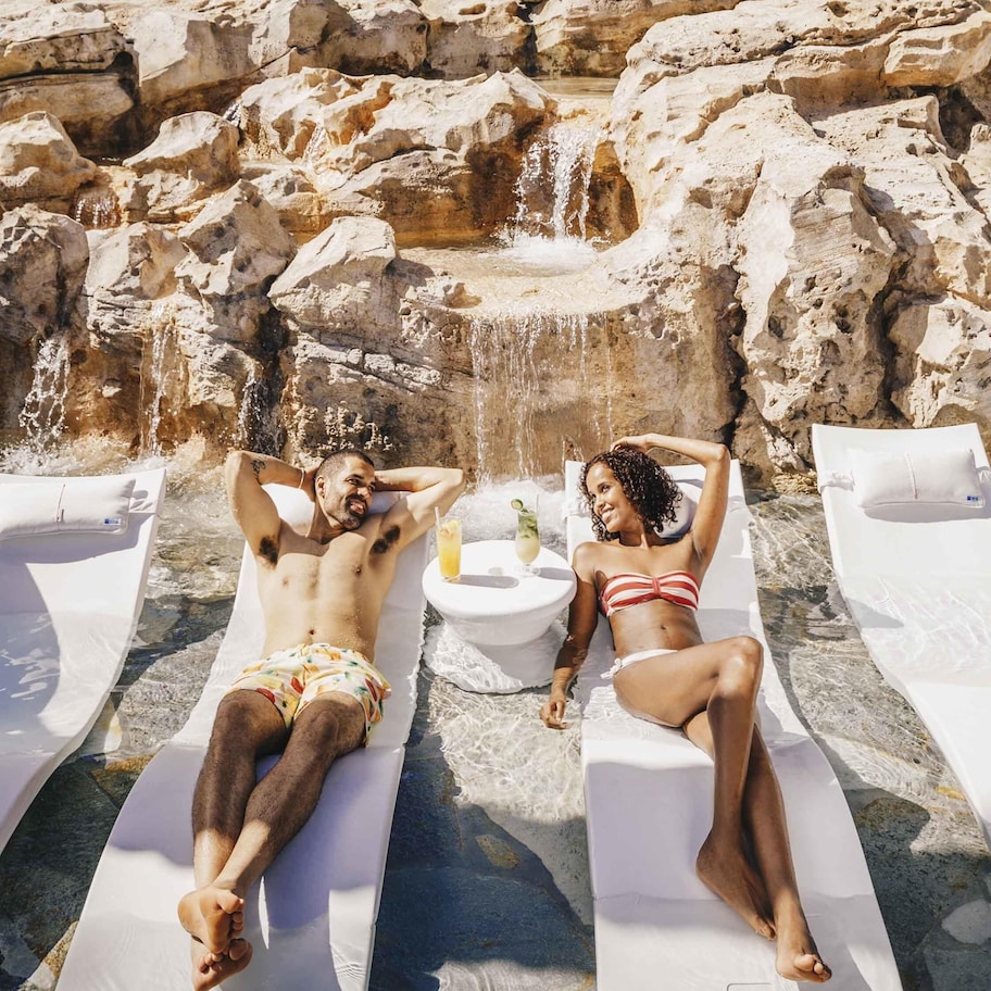 Two sun loungers with towels and a small table with a drink, set against a rocky backdrop with a waterfall.