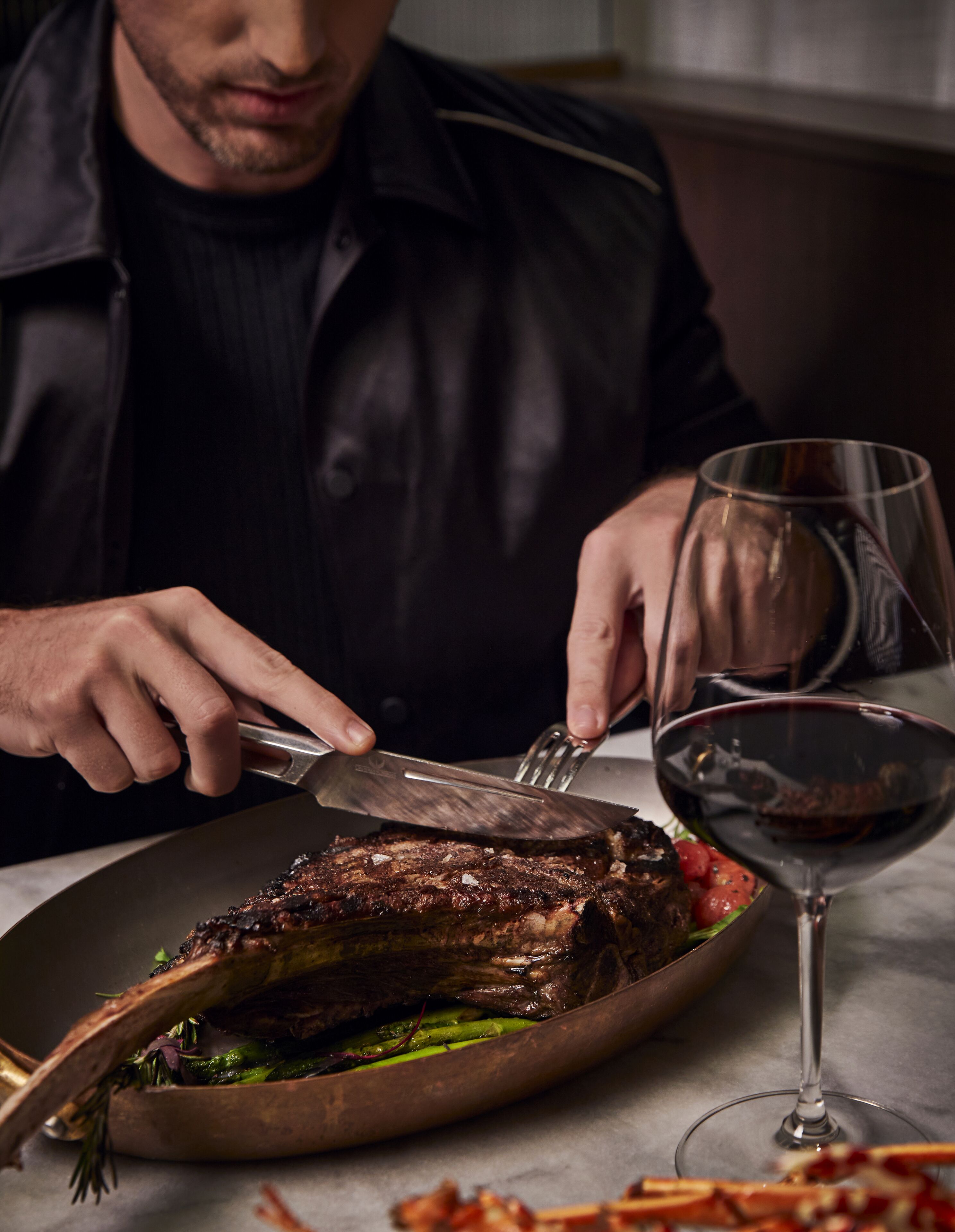 A person cutting steak with a fork and knife on a plate, with a glass of red wine and some vegetables on the table.