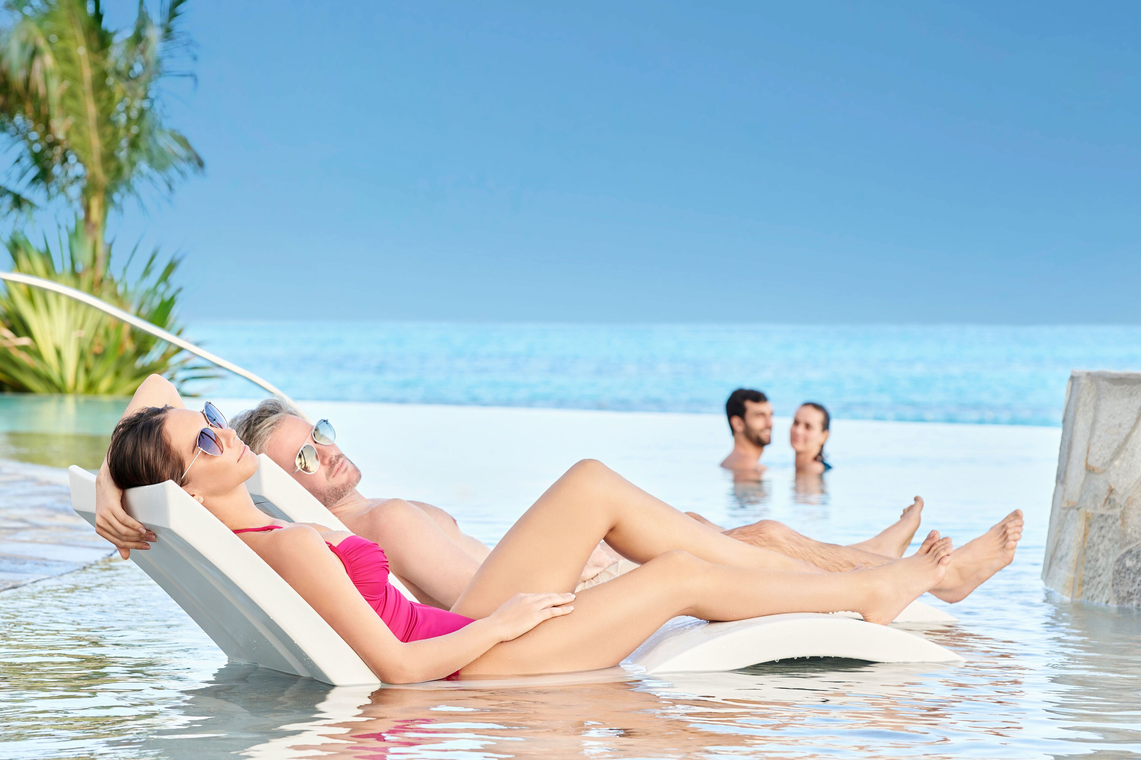 Un couple se détend sur des chaises longues au bord d’une piscine, avec une vue dégagée sur l’océan et des palmiers en arrière-plan.