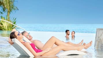 A couple relaxing on a lounge chair in a pool with a clear view of the ocean and palm trees in the background.