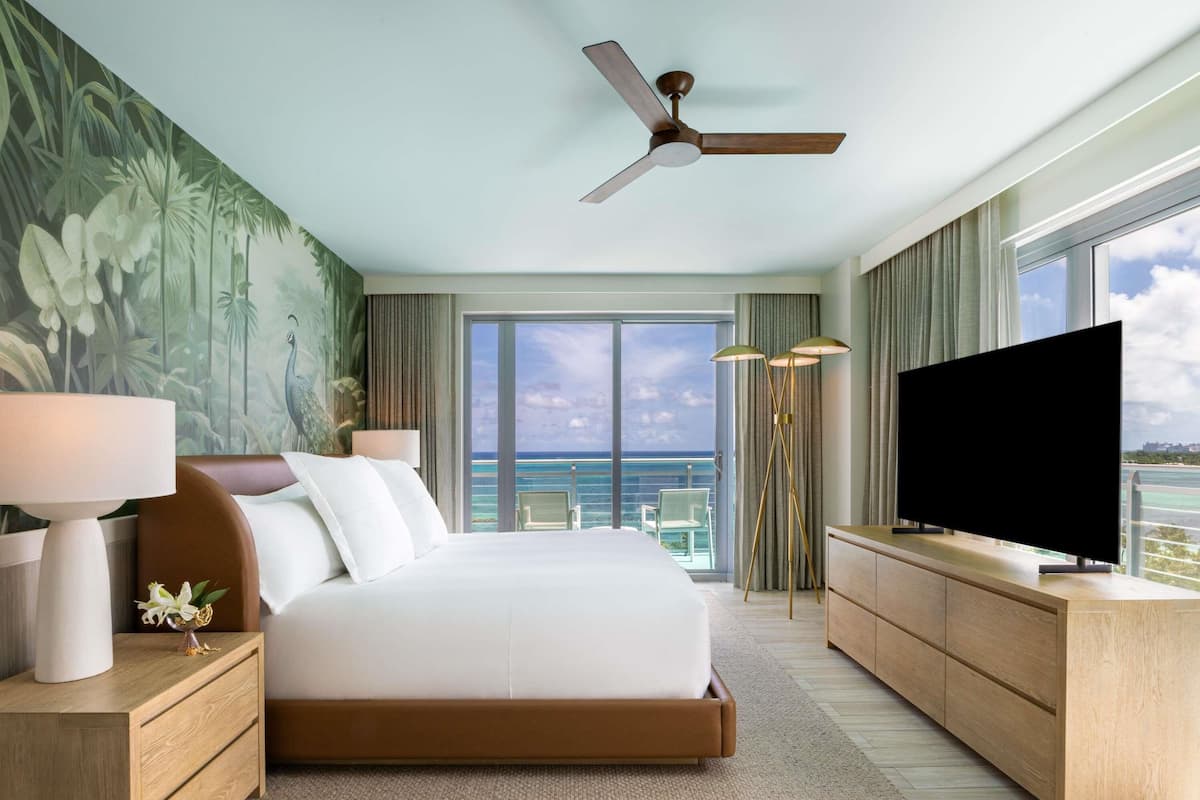 A hotel room with a bed, a TV, a ceiling fan, a large window with a view of the ocean, and a tropical-themed wall mural.