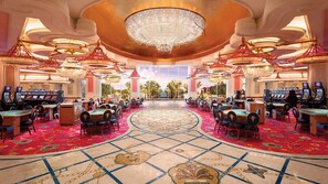 A casino with multiple gaming tables and chairs, a large chandelier, and a view of palm trees through large windows.
