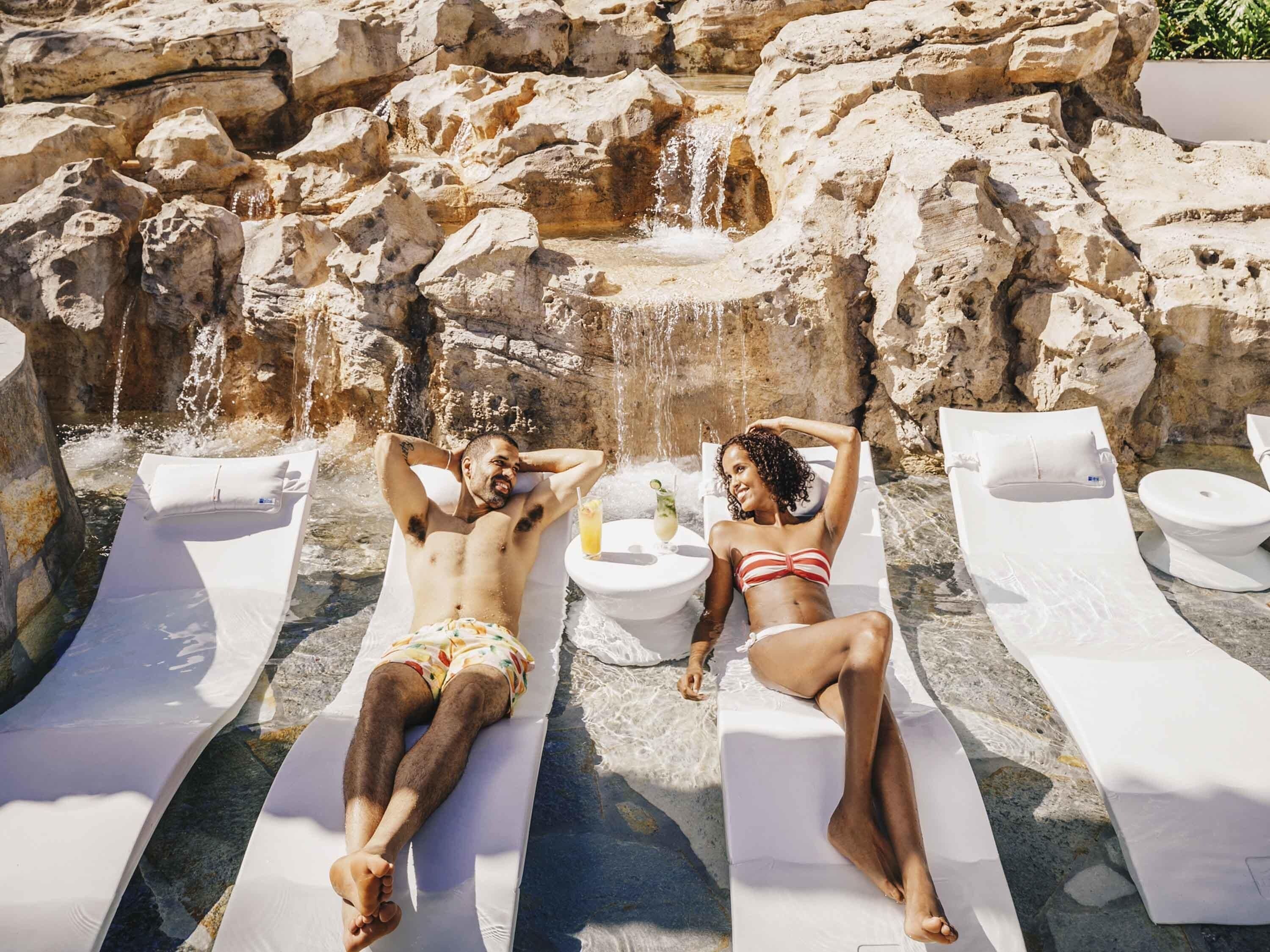 Deux chaises longues avec des serviettes et une petite table sur laquelle se trouve une boisson, le tout installé devant un paysage rocheux avec une cascade.