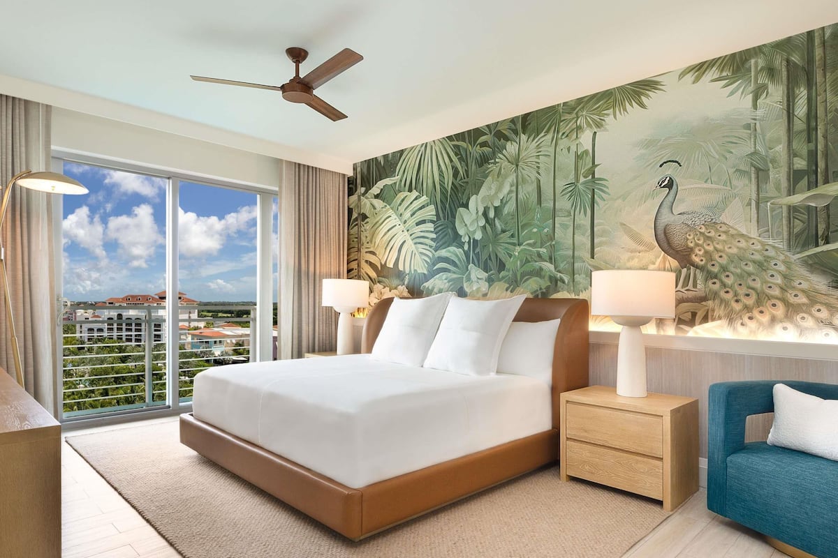 A hotel room with a large bed, two bedside tables with lamps, a blue armchair, a peacock mural, and a city view through the window.