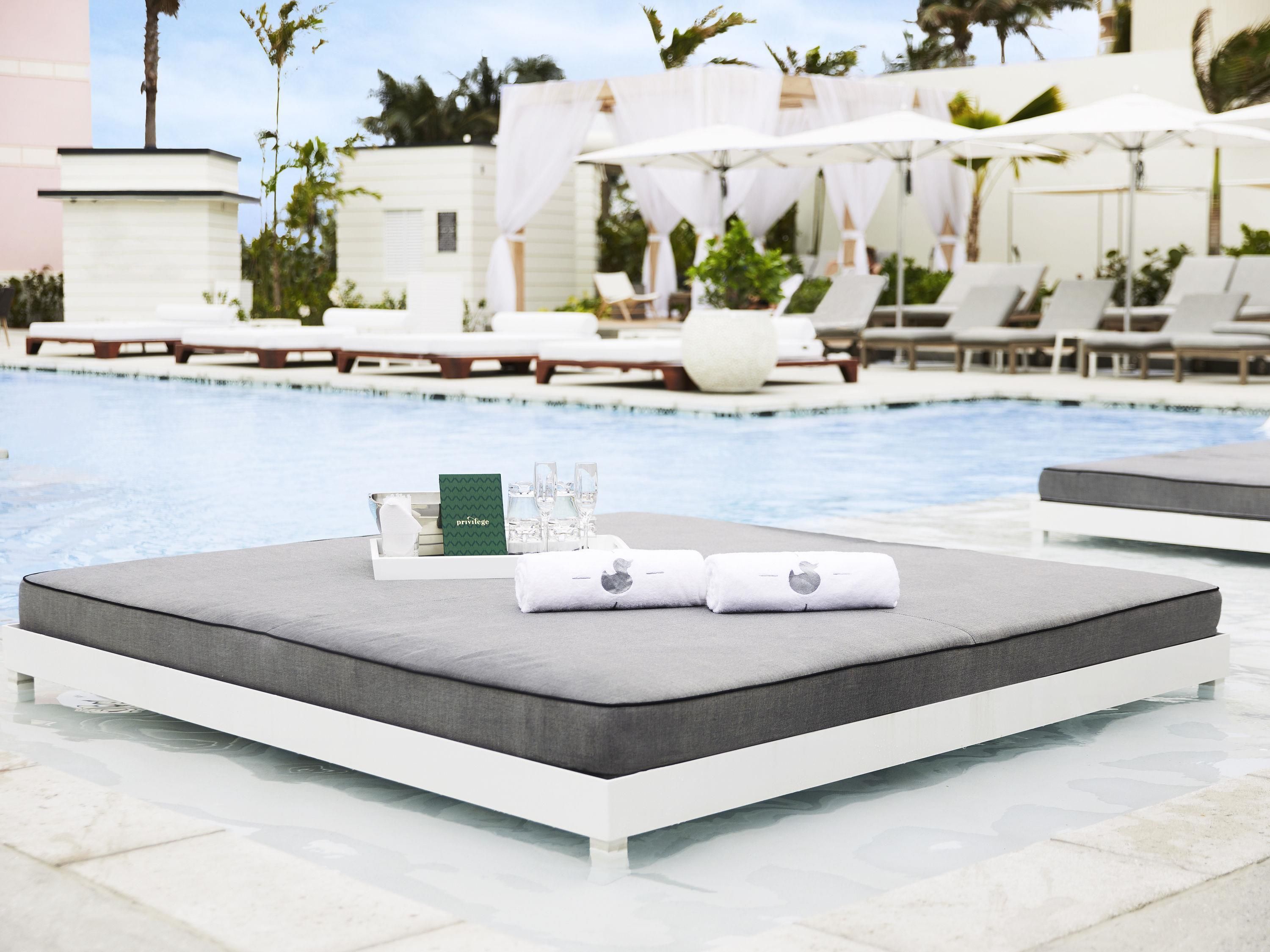 A poolside lounge area with a white lounge chair, a tray with a towel, a glass of water, and a card.