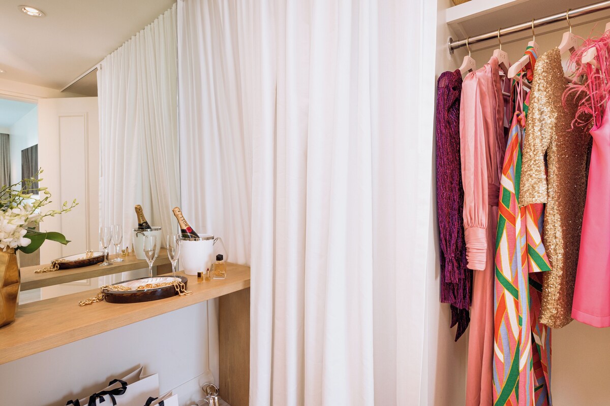 A bathroom with a vanity, a mirror, a shower curtain, and a rack of colorful dresses.