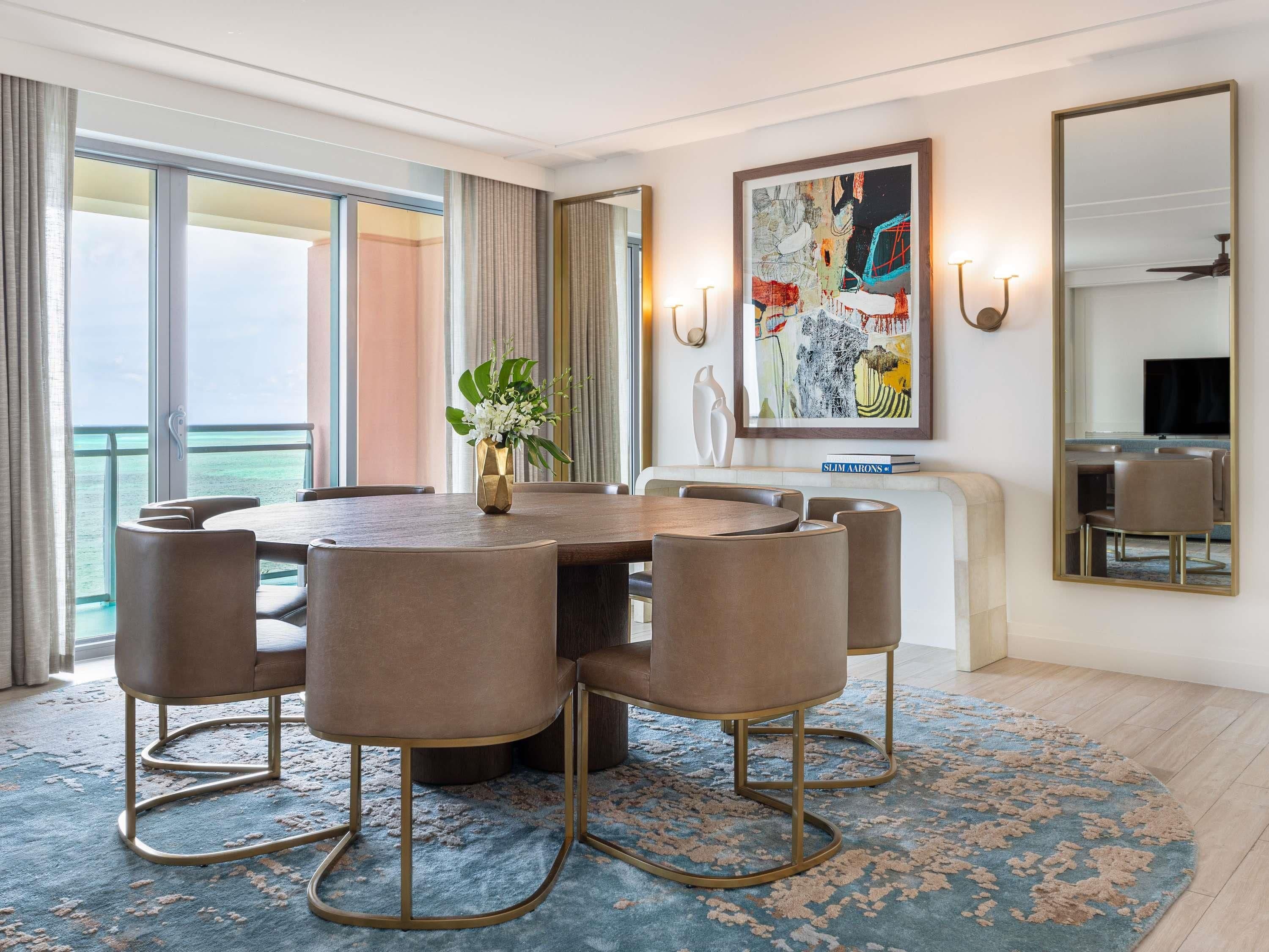 A dining area with a round table, six chairs, a vase with flowers, and a large abstract painting on the wall.