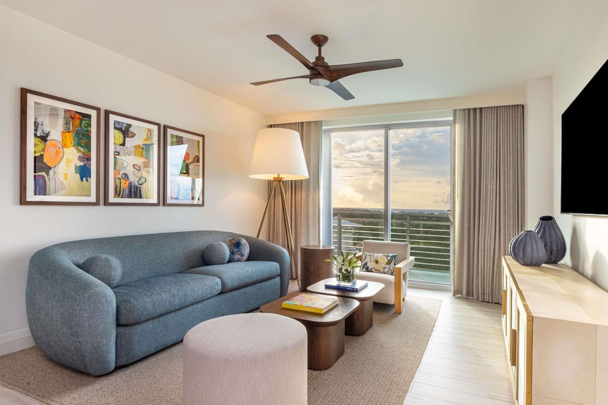 A modern hotel room with a sofa, ottoman, coffee table, and a TV. There are framed artworks on the wall, a ceiling fan, and a large window with a view.