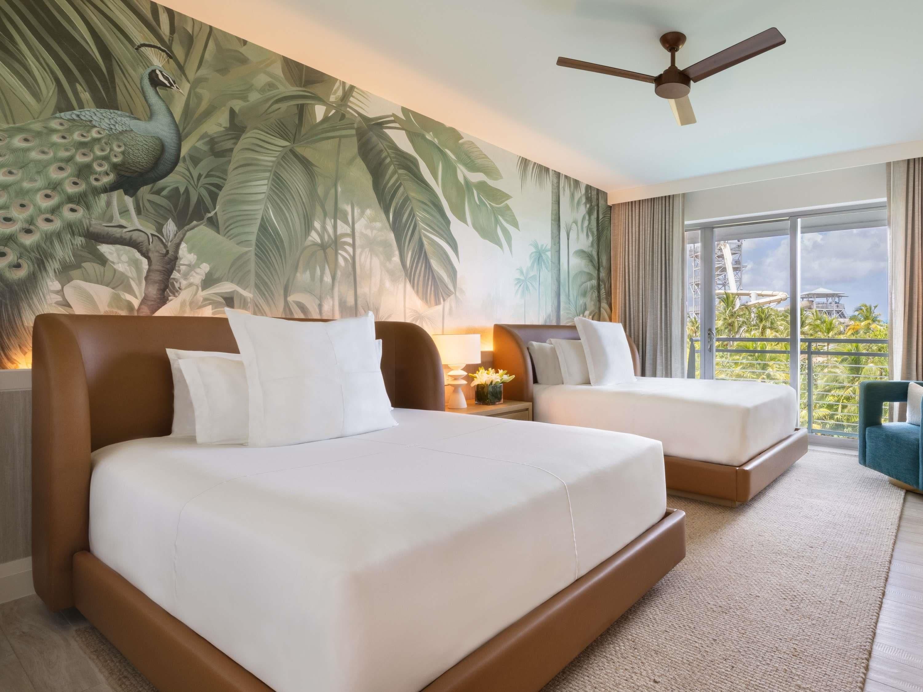 A hotel room with two beds, a ceiling fan, a large window with a view of palm trees, and a tropical mural on the wall.