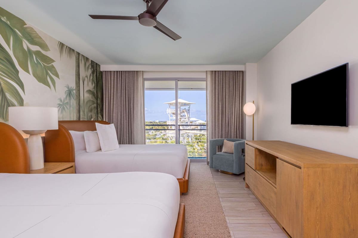 A hotel room with two beds, a ceiling fan, a TV, a desk, and a view of the ocean.