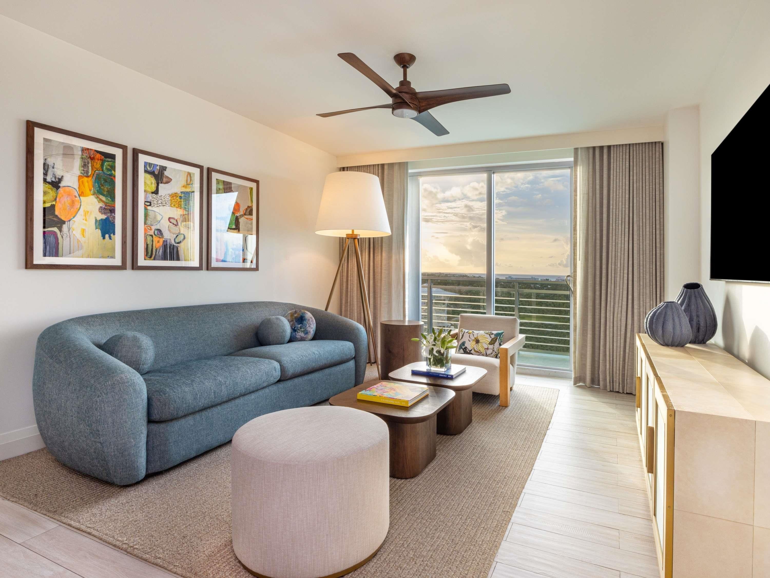 A modern hotel room with a sofa, ottoman, coffee table, and a TV. There are framed artworks on the wall, a ceiling fan, and a large window with a view.