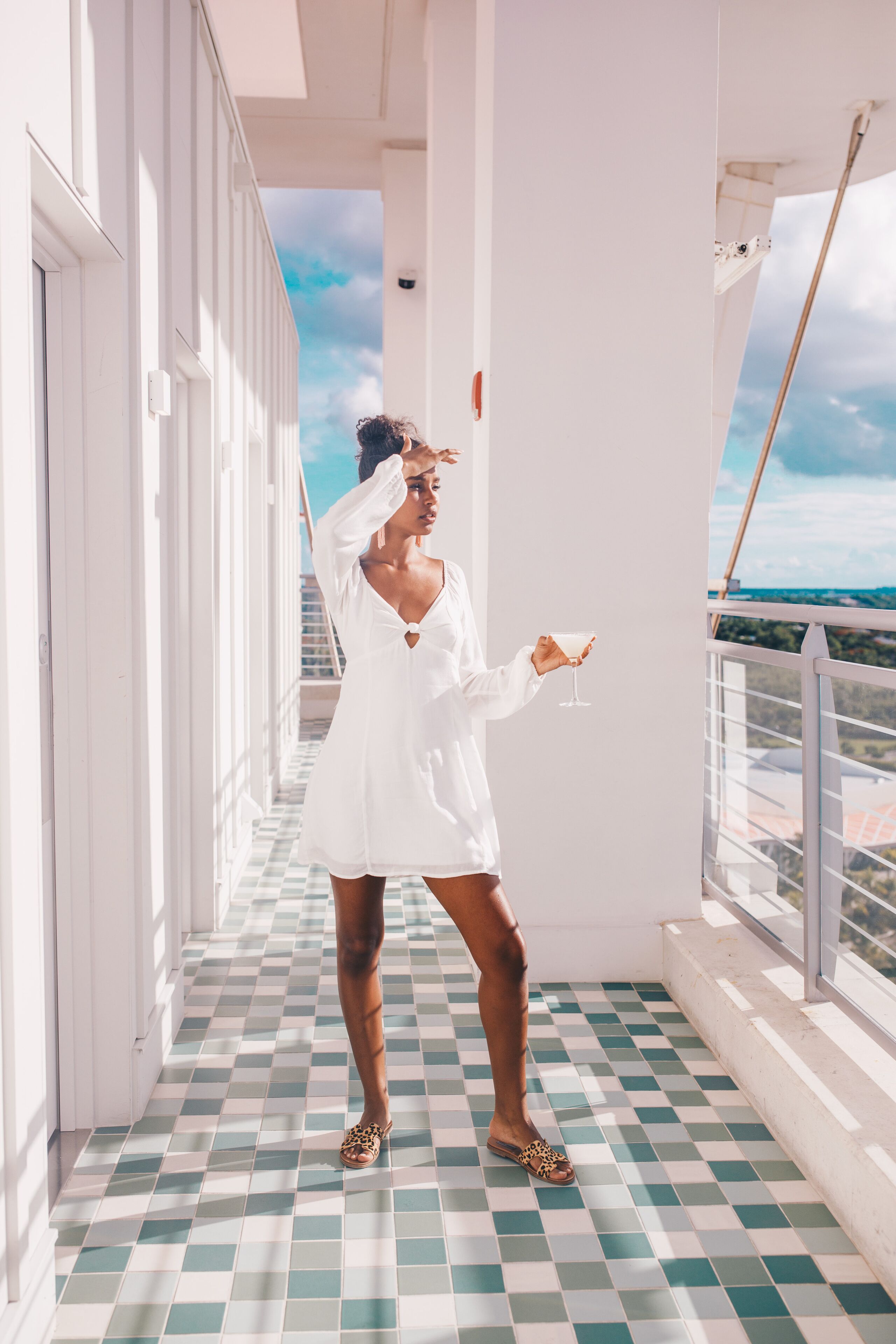 A woman in a white dress on a balcony with a checkered floor, holding a glass.