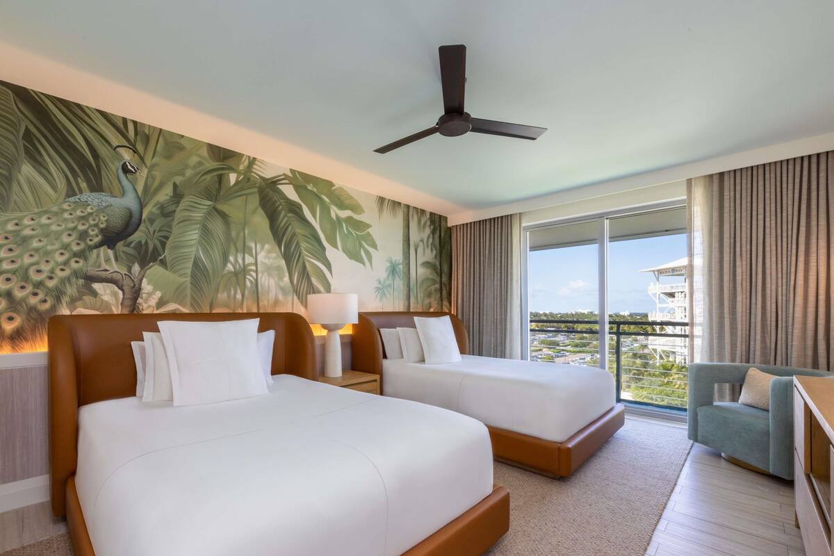 A hotel room with two beds, a peacock mural, a ceiling fan, and a balcony with a view.