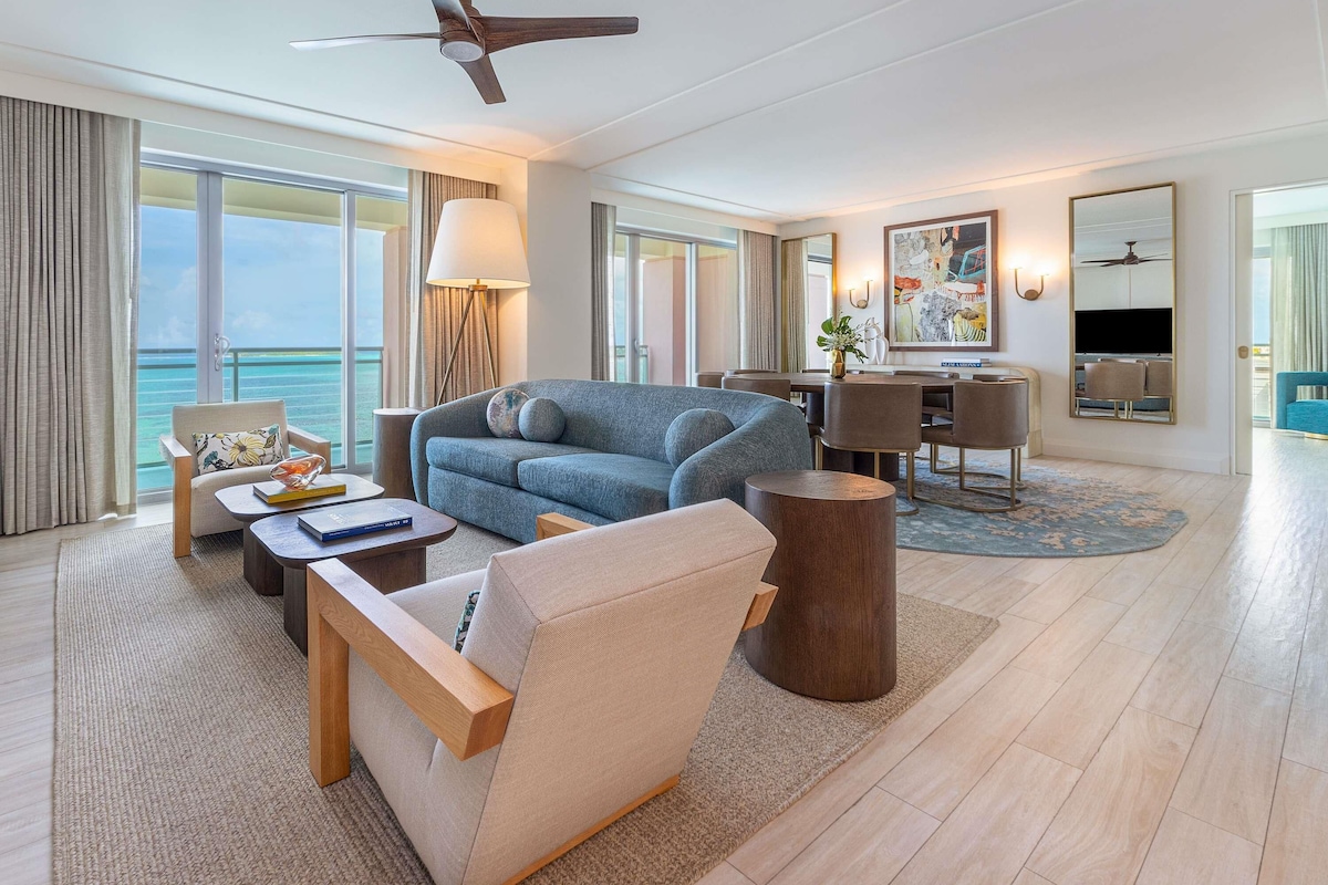 A spacious living room with a large sofa, a coffee table, and a wooden side table. There is a dining area with a table and chairs, and a large window offering a view of the ocean.