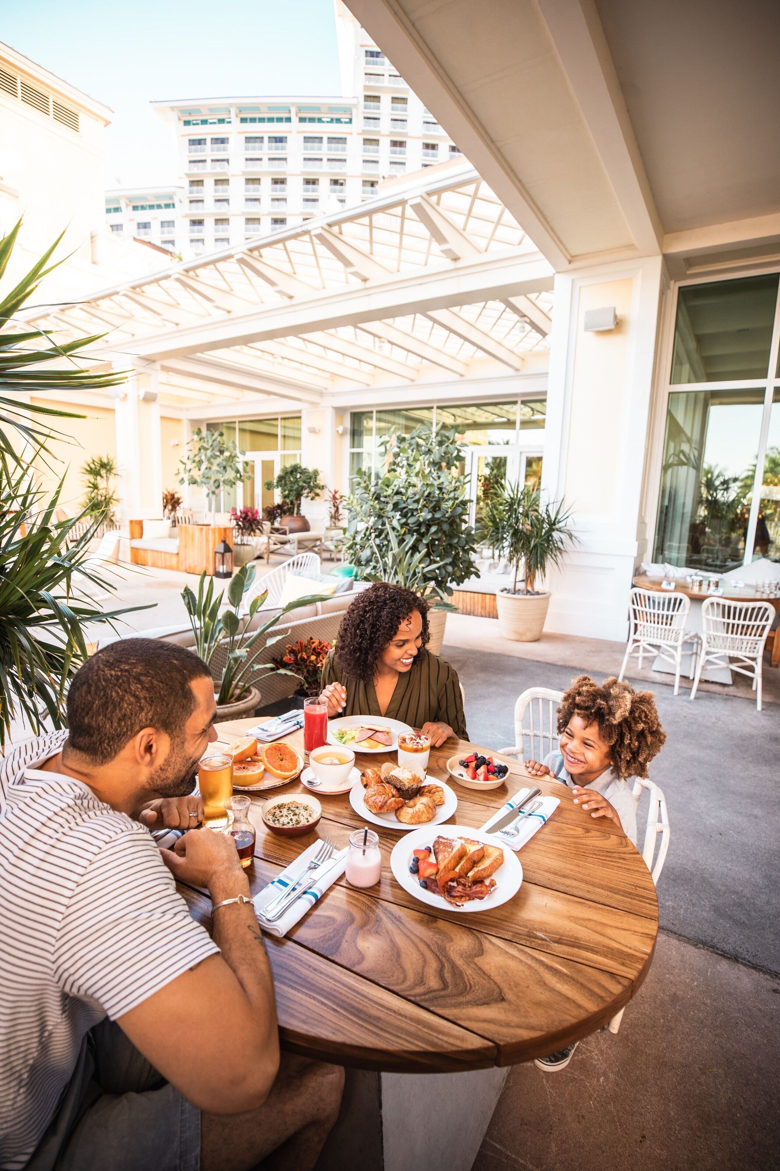 A family dining at an outdoor patio with a wooden table, surrounded by potted plants and modern architecture.