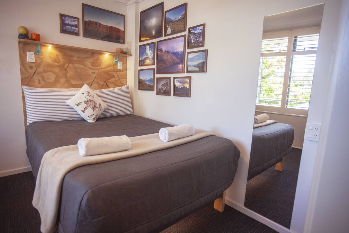 A hotel room with two beds, a wooden headboard, multiple framed pictures on the wall, and a window with blinds.