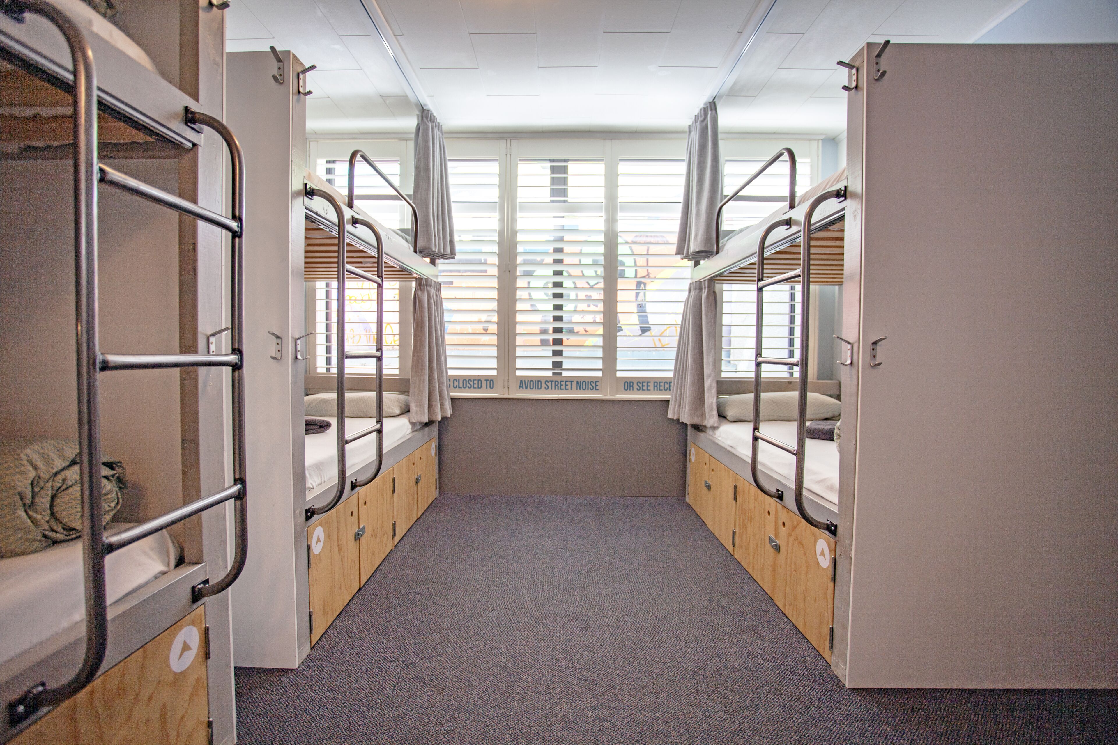 A dormitory room with bunk beds, a window with blinds, and a carpeted floor.