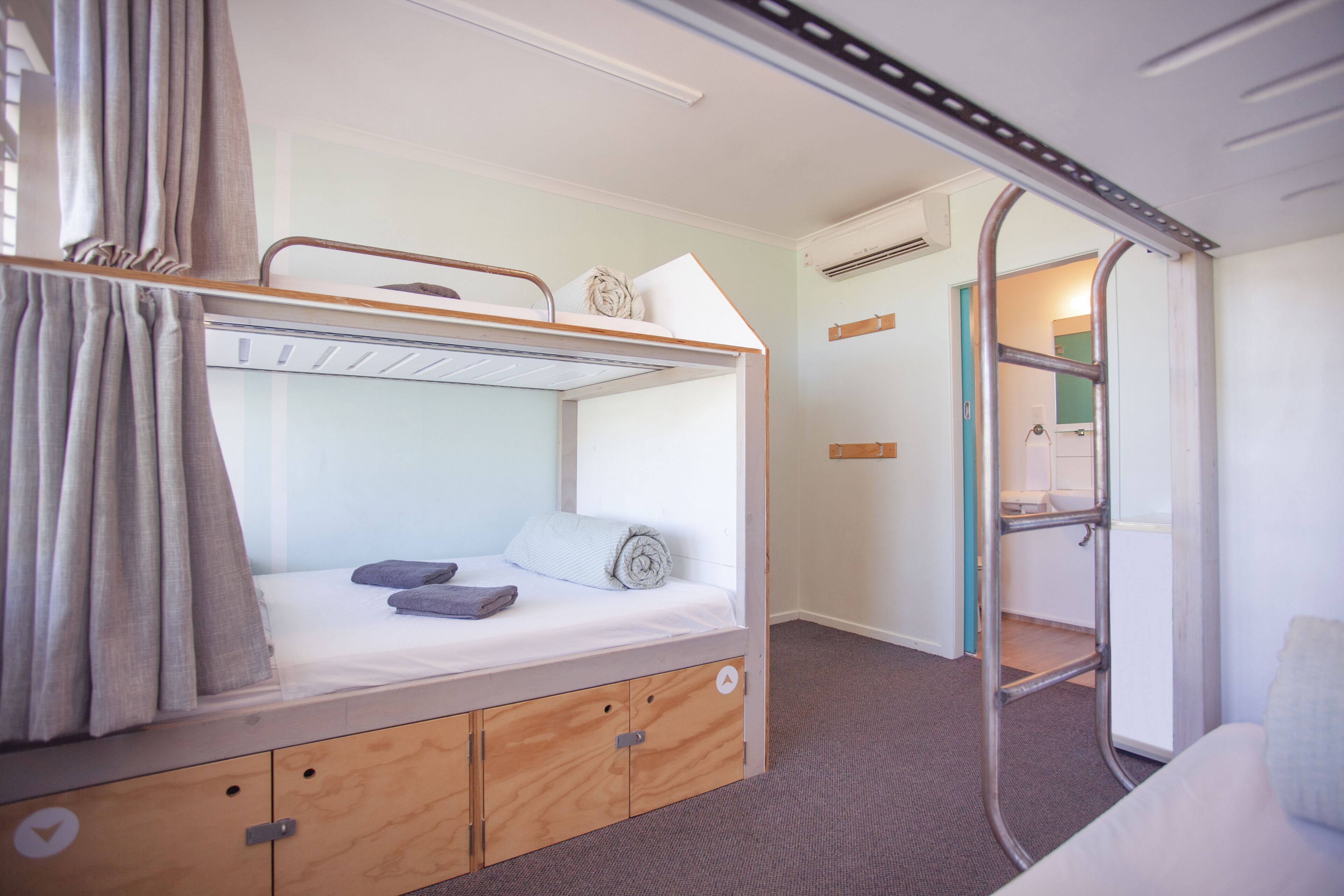 A bunk bed with a wooden lower bunk and a metal upper bunk. There is a small shelf on the lower bunk and a window with curtains.