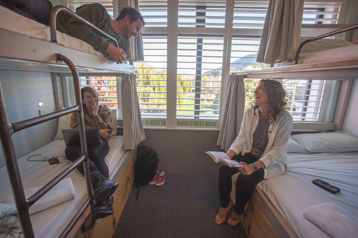 A dormitory room with bunk beds, a window with a view of greenery, and a closed window with a sign.