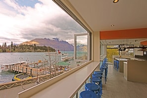 A modern interior with a view of a lake and mountains. It includes a bar area with stools and a kitchen counter. - Absoloot Hostel Queenstown (Queenstown)