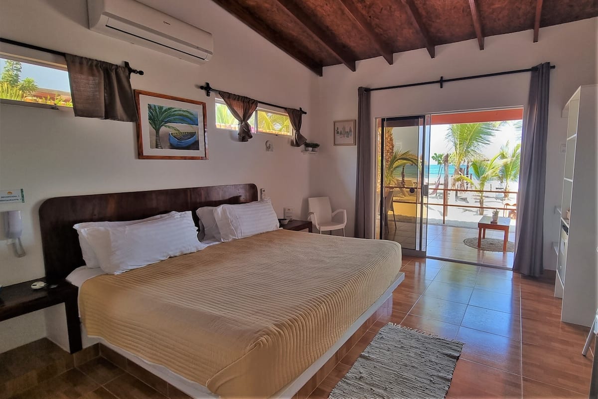 Ein Schlafzimmer mit einem großen Bett, einem Nachttisch, einem Schreibtisch und Blick auf den Strand durch eine Glasschiebetür.