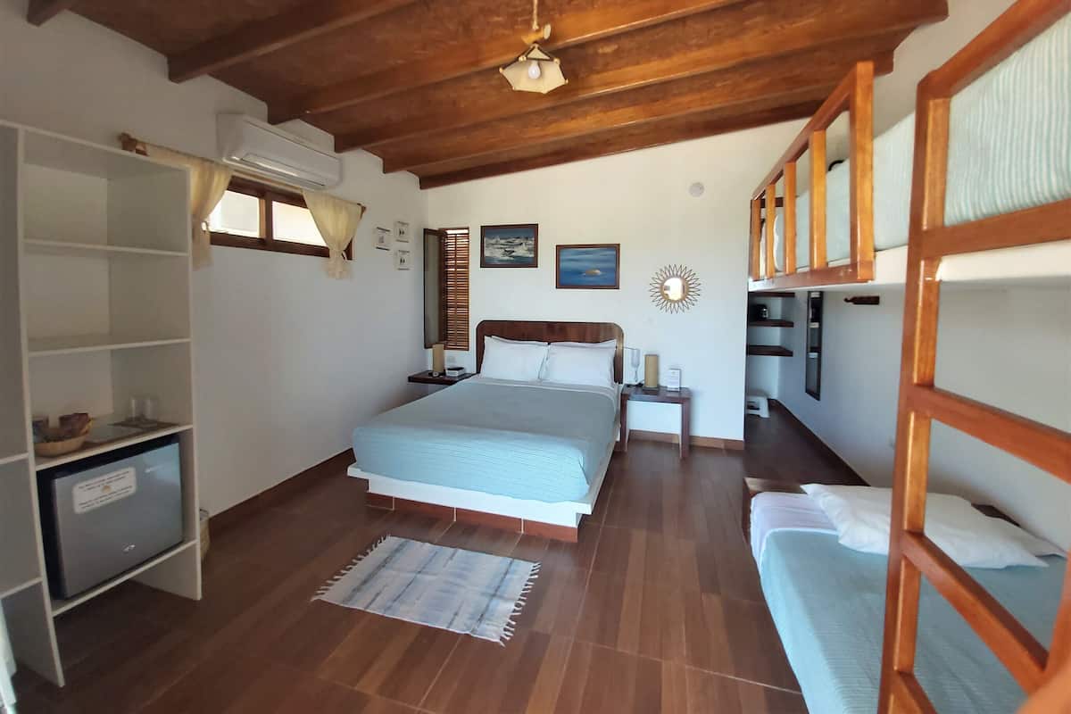 Ein Schlafzimmer mit Holzboden, einem Bett mit blauer Bettdecke, einem Nachttisch, einem Regal mit einem kleinen Gerät und einem Fenster mit Vorhängen.